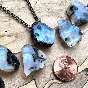 May include: A silver chain necklace with a raw, iridescent moonstone pendant. The pendant is a large, irregular shape with black inclusions. The moonstone is surrounded by smaller, raw moonstones of similar shape and size. A US penny is included for scale.