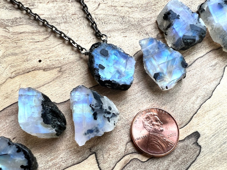 May include: A silver chain necklace with a raw, iridescent moonstone pendant. The pendant is a large, irregular shape with black inclusions. The moonstone is surrounded by smaller, raw moonstones of similar shape and size. A US penny is included for scale.