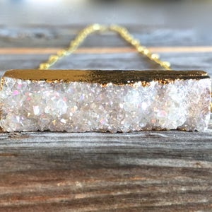 May include: A gold chain necklace with a white crystal bar pendant. The crystal is a cluster of small, iridescent crystals.