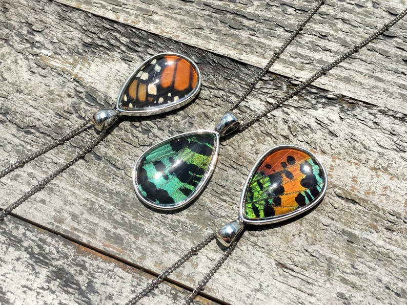 Peut inclure: Trois pendentifs en argent en forme de goutte d'eau avec des ailes de papillon ench&acirc;ss&eacute;es dans de la r&eacute;sine. Les pendentifs sont sur des cha&icirc;nes en argent. Les ailes de papillon sont orange, vertes et noires.