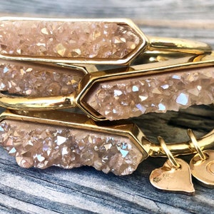May include: Three gold bangles with a light brown, crystal-like stone set in each. The bangles are stacked on top of each other. The bottom bangle has a gold heart charm and a gold charm with the letter "C" on it.