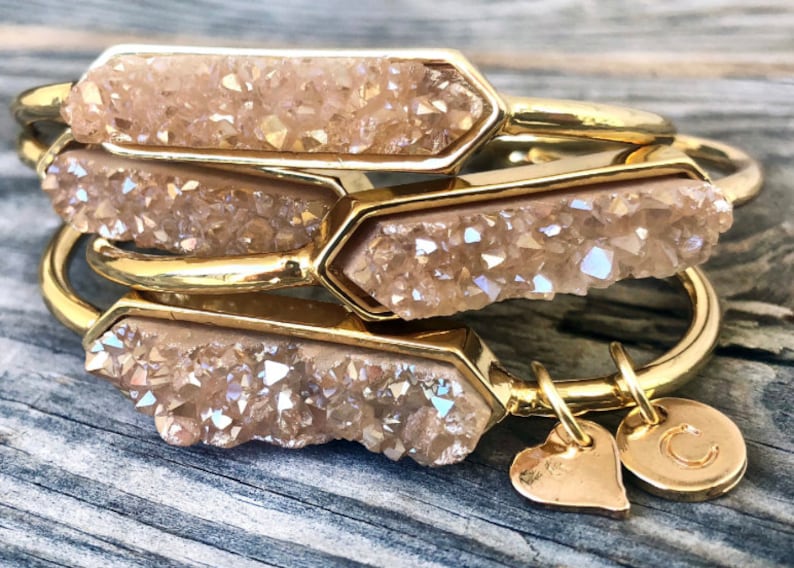 May include: Three gold bangles with a light brown, crystal-like stone set in each. The bangles are stacked on top of each other. The bottom bangle has a gold heart charm and a gold charm with the letter "C" on it.