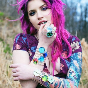 May include: A woman with pink hair wearing a purple floral dress and multiple silver bracelets with colourful glass insets. The bracelets are stacked on her left wrist.