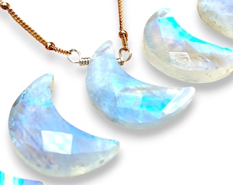 May include: A necklace featuring a delicate gold chain with a faceted, crescent moon-shaped pendant made of iridescent, translucent stone. The stone displays blue and white light reflections, and the pendant is attached to the chain with a wire loop.