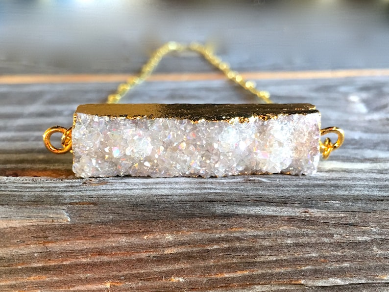 May include: A gold chain necklace with a white crystal bar pendant. The crystal is a cluster of small, white crystals with a gold rim.