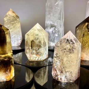 May include: A collection of seven clear and golden yellow crystal points, some with a smoky brown hue, arranged on black circular bases. The crystals are all pointed and have a natural, rough texture.