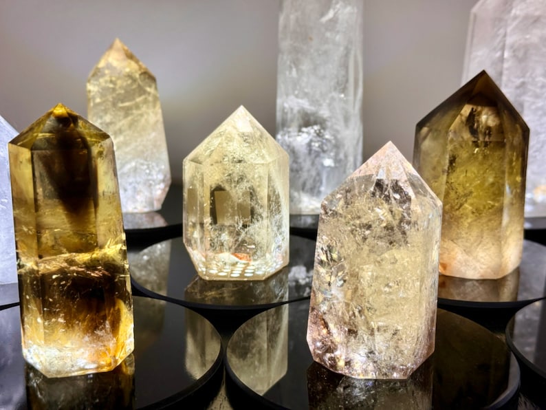 May include: A collection of seven clear and golden yellow crystal points, some with a smoky brown hue, arranged on black circular bases. The crystals are all pointed and have a natural, rough texture.