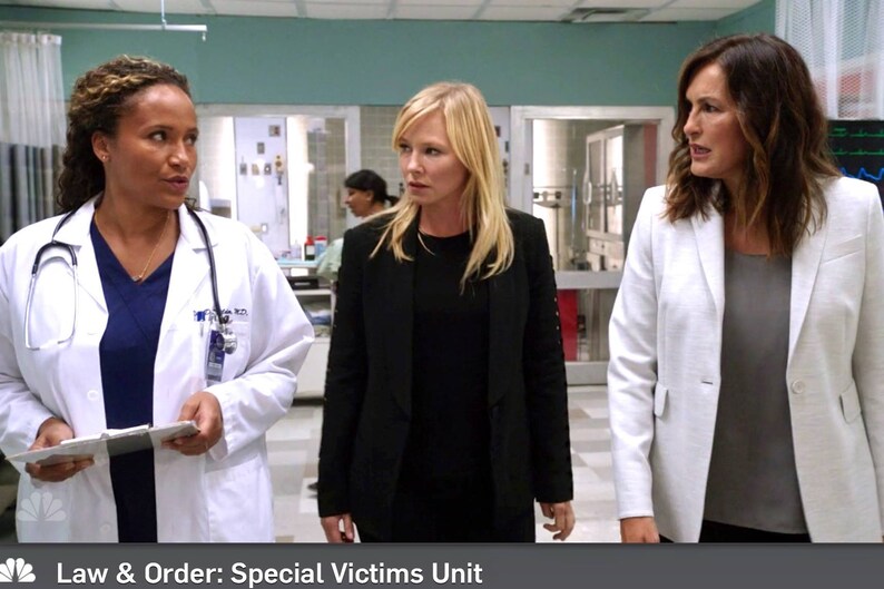 May include: Three women in a hospital setting. The woman on the left is wearing a white lab coat over a blue shirt and a stethoscope around her neck. The woman in the middle is wearing a black blazer over a black shirt. The woman on the right is wearing a white blazer over a gray shirt. The text "Law & Order: Special Victims Unit" is at the bottom of the image.