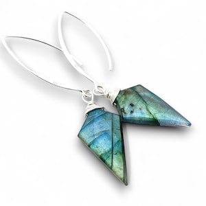 May include: A pair of silver drop earrings featuring faceted, triangular labradorite stones. The stones display iridescent blue and green hues, and are wire-wrapped to the silver hooks. The earrings are set against a white background.