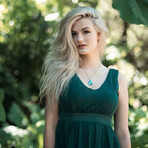 Peut inclure: Une femme aux longs cheveux blonds portant une robe sans manches vert fonc&eacute; avec un collier &agrave; pendentif en forme de larme.