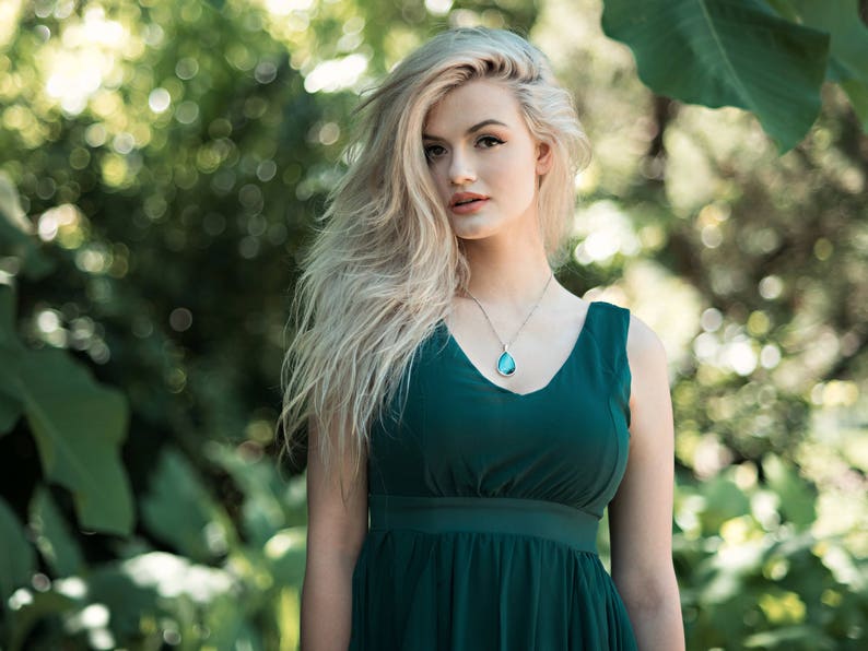 Peut inclure: Une femme aux longs cheveux blonds portant une robe sans manches vert fonc&eacute; avec un collier &agrave; pendentif en forme de larme.