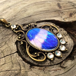 Op de afbeelding: Een vintage-stijl hangerketting met een ovale, iriserende blauw-paarse middensteen. De steen is gezet in een bronskleurig metalen frame met sierlijke krullen en heldere kristallen accenten. De ketting is ook bronskleurig.