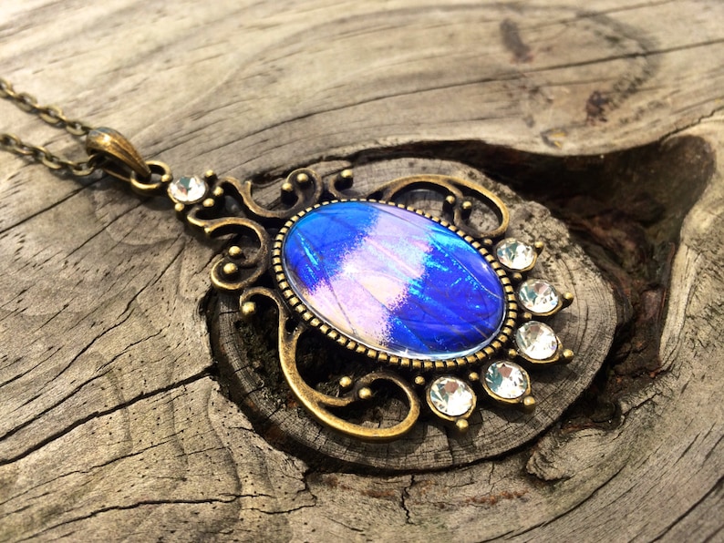 Op de afbeelding: Een vintage-stijl hangerketting met een ovale, iriserende blauw-paarse middensteen. De steen is gezet in een bronskleurig metalen frame met sierlijke krullen en heldere kristallen accenten. De ketting is ook bronskleurig.