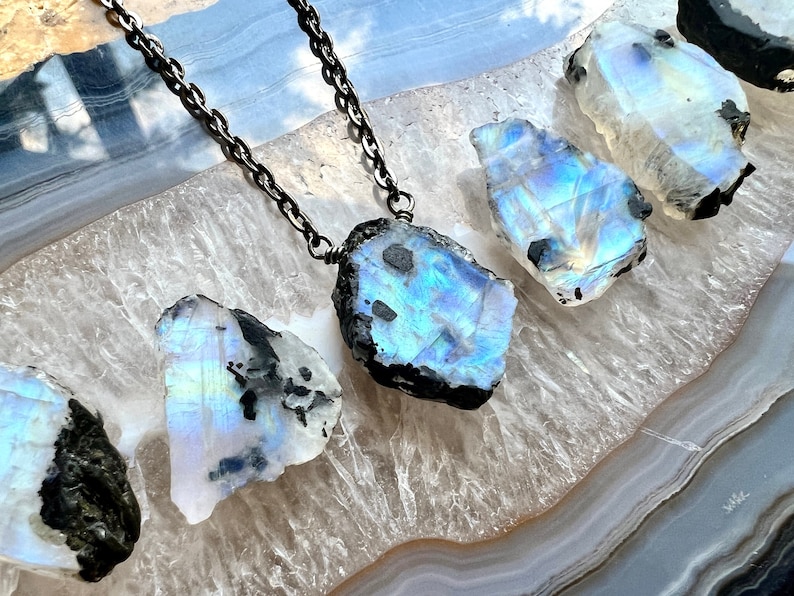 May include: A silver chain necklace with a large, iridescent, blue and white moonstone pendant. The pendant is raw and has a black, rough exterior. The necklace is displayed on a white and gray agate slice.
