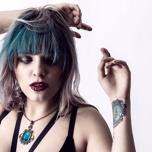 May include: A woman with teal blue hair and a dark blue halter top is wearing a necklace with a large, oval, blue gemstone pendant. She has a tattoo on her wrist of a blue eye with a gold frame.