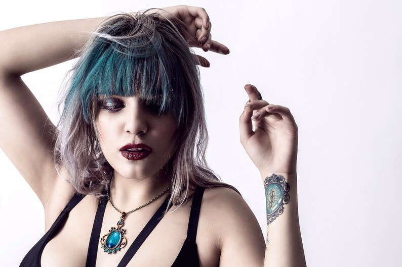 May include: A woman with teal blue hair and a dark blue halter top is wearing a necklace with a large, oval, blue gemstone pendant. She has a tattoo on her wrist of a blue eye with a gold frame.
