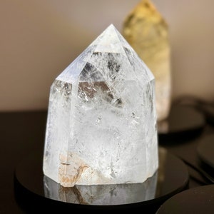 May include: A clear quartz crystal point, also known as a crystal wand, is displayed on a black circular stand. The crystal is clear and has a slight iridescent quality. The crystal is a popular choice for meditation and healing.