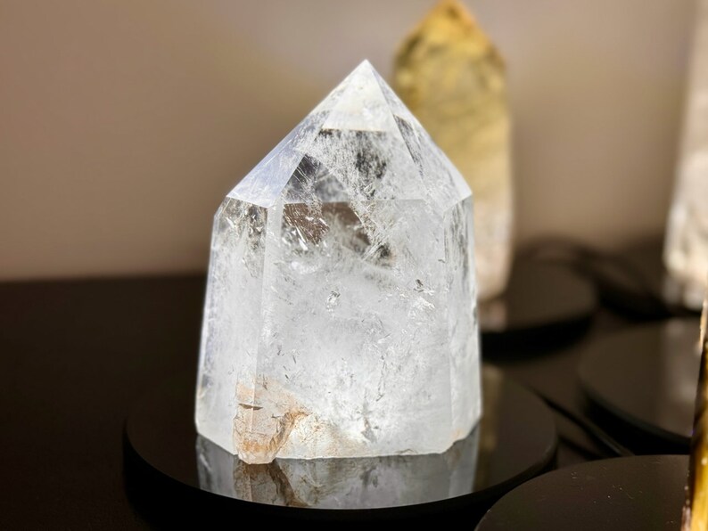 May include: A clear quartz crystal point, also known as a crystal wand, is displayed on a black circular stand. The crystal is clear and has a slight iridescent quality. The crystal is a popular choice for meditation and healing.