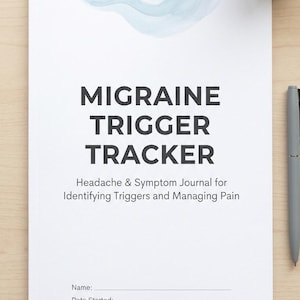 Op de afbeelding: Een wit journaal met de titel "MIGRAINE TRIGGER TRACKER" en de ondertitel "Headache & Symptom Journal for Identifying Triggers and Managing Pain." Een vetplant in een witte pot en een grijze pen zijn ook zichtbaar.