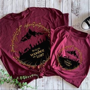 May include: Two maroon t-shirts with a gold ring design and the text "Not all who wander are lost." Black and white trainers are next to the shirts. The shirts have a mountain and silhouette design.