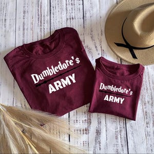 May include: Two maroon t-shirts with white text that reads "Dumbledore's Army" and a graphic of a wand.