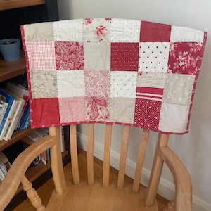 May include: A red and white patchwork quilt with a floral pattern is draped over the back of a wooden chair.