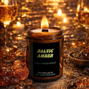 Könnte beinhalten: Eine brennende, bernsteinfarbene Kerze in einem Glas mit einem schwarzen Etikett mit der Aufschrift "Baltic Amber, Forest Woods Scents". Die Kerze ist von bernsteinfarbenen Kristallen und einem verschwommenen Hintergrund aus Kerzen und Gläsern umgeben.