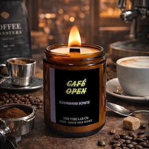 Könnte beinhalten: Eine brennende, braune Glas-Kerze mit einem schwarzen Etikett mit der Aufschrift "Café Open" in Gelb. Die Kerze ist von Kaffeebohnen, einer Tasse Espresso und einer Tasse Cappuccino umgeben. Die Kerze ist von The Vibe Lab Co.