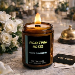Könnte beinhalten: Eine brennende Kerze in einem braunen Glas mit einem schwarzen Etikett mit der Aufschrift "SIGNATURE HOTEL, FRESH & CLEAN SCENTS". Neben der Kerze befinden sich eine Hotelglocke und Karten mit der Aufschrift "THE VIBE".