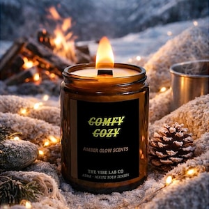 Könnte beinhalten: Eine brennende, bernsteinfarbene Kerze in einem Glas mit einem schwarzen Etikett mit der Aufschrift "COMFY COZY" in goldfarbenen Buchstaben. Die Kerze ist von einer verschneiten Landschaft, einem Tannenzapfen und einem Lagerfeuer im Hintergrund umgeben.