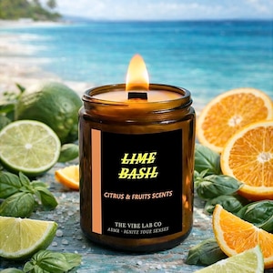 Könnte beinhalten: Eine brennende Kerze in einem braunen Glas mit einem schwarzen Etikett mit der Aufschrift "LIME BASIL" und "CITRUS & FRUITS SCENTS". Die Kerze ist von Limettenscheiben, Orangenscheiben und Basilikumblättern umgeben. Der Hintergrund ist eine verschwommene Strandszene.