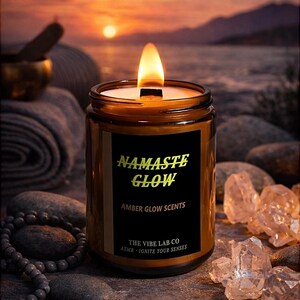 Könnte beinhalten: Eine brennende, bernsteinfarbene Kerze in einem Glas mit einem schwarzen Etikett mit der goldenen Aufschrift "Namaste Glow". Die Kerze ist von Felsen und Kristallen umgeben, mit einem Sonnenuntergang im Hintergrund. Das Etikett lautet auch "Amber Glow Scents" und "The Vibe Lab Co".