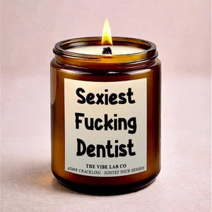 Puede incluir: Una vela encendida en un tarro de cristal marrón con una etiqueta blanca que dice "Sexiest Fucking Dentist" en texto negro. La vela es de The Vibe Lab Co. y tiene crepitación ASMR. La vela tiene una mecha encendida.