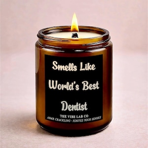 Puede incluir: Una vela encendida en un frasco de vidrio marrón con una etiqueta negra. La etiqueta dice "Smells Like World's Best Dentist" en escritura blanca. La vela es de The Vibe Lab Co.