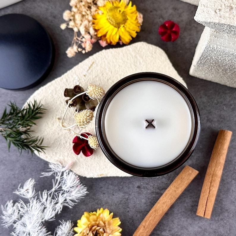 May include: A top-down view of a lit candle in a dark brown glass container. The candle has a white wax surface with a wooden wick. The candle is surrounded by dried flowers, cinnamon sticks, and a black lid on a textured surface.
