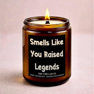 May include: A lit brown glass jar candle with a black label that reads "Smells Like You Raised Legends." The candle has a white wax and a burning wick. The text "THE VIBE LAB CO" and "ASMR CRACKLING - IGNITES YOUR SENSES" are also on the label.