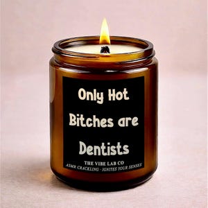 May include: A lit candle in a brown glass jar with a black label. The label reads "Only Hot Bitches are Dentists" in white text. The bottom of the label says "THE VIBE LAB CO ASMR CRACKLING - IGNITES YOUR SENSES".