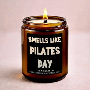 May include: A lit candle in a brown glass jar. The black label reads "Smells Like Pilates Day" in white text. The bottom of the label says "The Vibe Lab Co" and "ASMR Crackling - Ignites Your Senses". The candle has a white wax and a yellow flame.