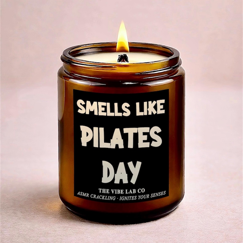 May include: A lit candle in a brown glass jar. The black label reads "Smells Like Pilates Day" in white text. The bottom of the label says "The Vibe Lab Co" and "ASMR Crackling - Ignites Your Senses". The candle has a white wax and a yellow flame.