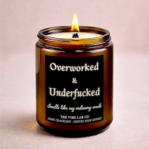 May include: A lit brown glass jar candle with a black label. The label reads "Overworked & Underfucked" with the text "Smells like my ordinary week" and "The Vibe Lab Co". The candle has a lit wick.
