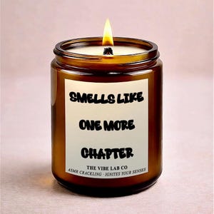 Op de afbeelding: Een brandende bruine glazen kaarsenpot met een wit etiket met de tekst "Smells Like One More Chapter". De kaars heeft een brandende lont en de tekst "The Vibe Lab Co" en "ASMR Crackling - Ignites Your Senses" onderaan het etiket.