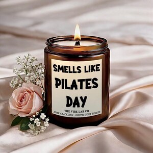 May include: A lit brown glass candle jar with the text "Smells Like Pilates Day" on a white label. A pink rose and white flowers are to the left of the candle. The candle is on a satin surface.