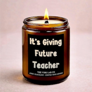 May include: A lit candle in a brown glass jar. The black label reads "It's Giving Future Teacher" in white text. The bottom of the label says "The Vibe Lab Co" and "ASMR CRACKLING - IGNITES YOUR SENSES".