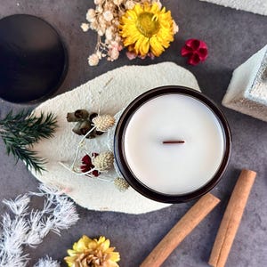 May include: A candle in a dark brown glass jar with a white wax interior and a wooden wick. The candle is surrounded by dried flowers, cinnamon sticks, and a black lid on a textured white surface.