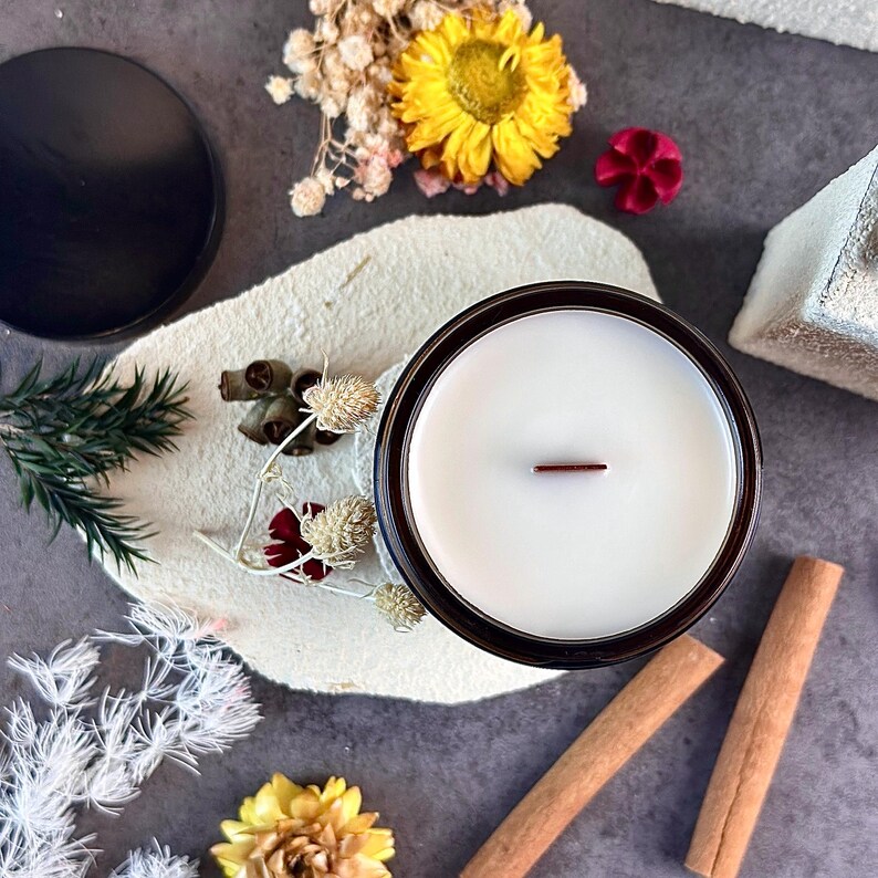 May include: A candle in a dark brown glass jar with a white wax interior and a wooden wick. The candle is surrounded by dried flowers, cinnamon sticks, and a black lid on a textured white surface.