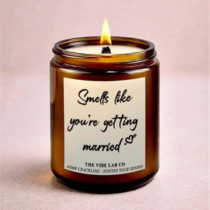 May include: A burning brown glass candle with a white label. The label says "Smells like you're getting married" with a heart and arrow graphic. The text "THE VIBE LAB CO ASMR CRACKLING - IGNITES YOUR SENSES" is also visible.