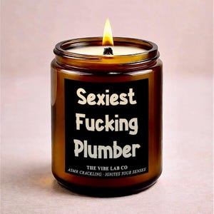 Puede incluir: Una vela encendida en un tarro de vidrio marrón con una etiqueta negra que dice "Sexiest Fucking Plumber" en texto blanco. La vela es de The Vibe Lab Co. y tiene crepitación ASMR.