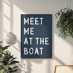 May include: A framed navy blue art print with the words "MEET ME AT THE BOAT" in white block letters. The print hangs on a white wall, with a wooden chair and potted plants in the foreground. A modern home decor item.