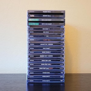 May include: A pile of PlayStation game cases, predominantly black with white text. Titles visible include "Resident Evil 2", "Metal Gear Solid", "Dino Crisis 2", "Silent Hill", and "Grand Theft Auto". The cases are stacked vertically.