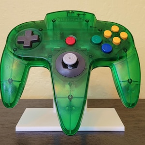May include: A clear green Nintendo 64 controller with gray buttons and a white base. The controller has a red button, a white button, and a gray joystick.
